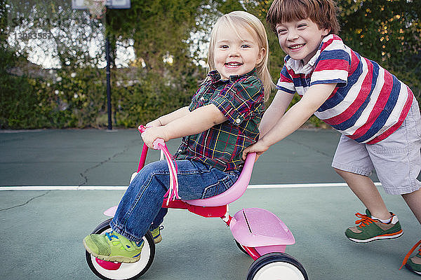 Porträt eines glücklichen Jungen  der seine Schwester schiebend auf einem Dreirad im Hof sitzt