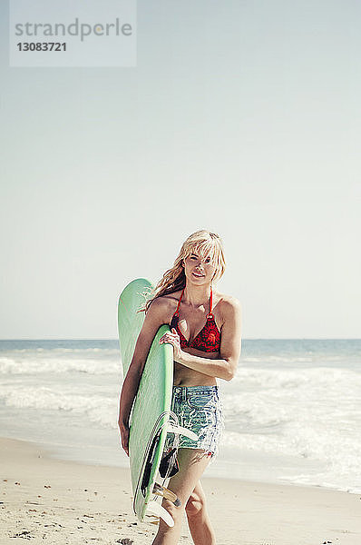 Porträt einer selbstbewussten Frau  die beim Strandspaziergang ein Surfbrett trägt