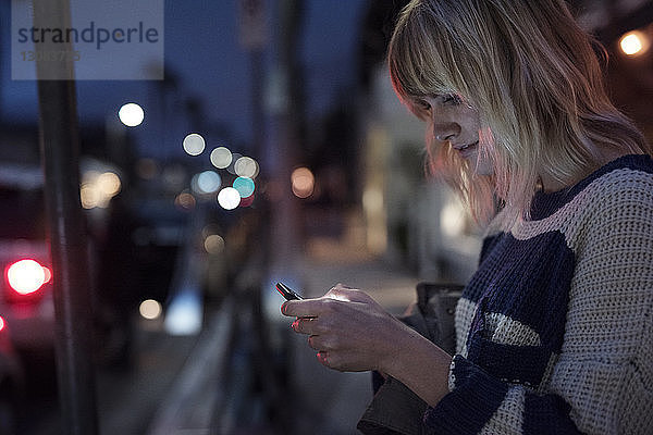 Seitenansicht einer Frau  die ein Smartphone in einer beleuchteten Stadt bei Nacht benutzt