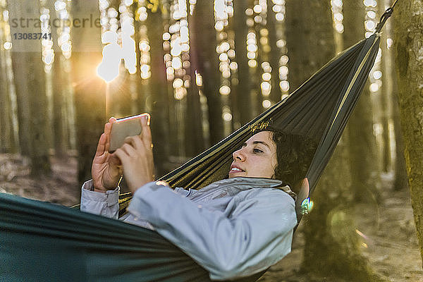 Frau benutzt Mobiltelefon  während sie bei Sonnenuntergang auf einer Hängematte im Wald liegt