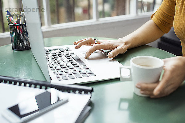 Beschnittenes Bild einer Geschäftsfrau  die eine Kaffeetasse hält  während sie im Büro einen Laptop benutzt