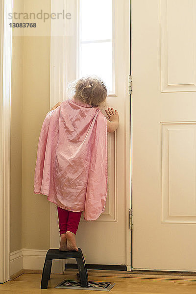 Rückansicht eines Mädchens mit Umhang  das zu Hause auf einem Hocker vor der Tür steht