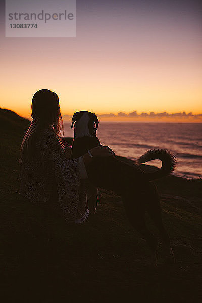 Seitenansicht einer Frau  die bei Sonnenuntergang mit Hund auf einem Hügel sitzt