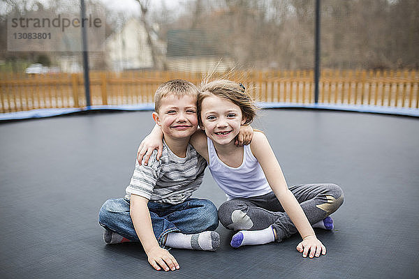 Porträt von glücklichen Geschwistern  die auf dem Spielplatz auf einem Trampolin sitzen