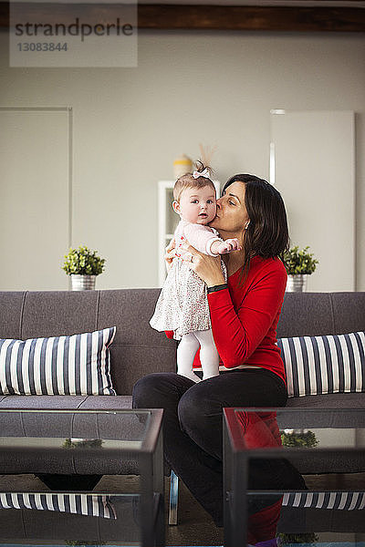 Mutter küsst ihre kleine Tochter  während sie zu Hause auf dem Sofa sitzt