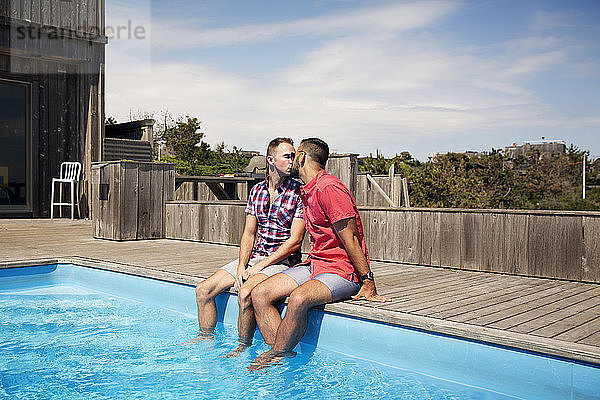 Homosexuelles Paar küsst sich  während es am Poolrand gegen den Himmel sitzt