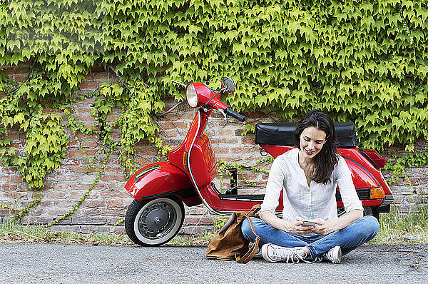 Frau benutzt Mobiltelefon  während sie gegen Motorroller und Efeuwand sitzt