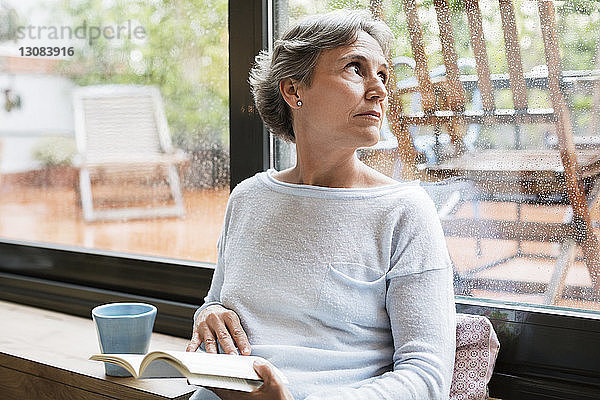 Nachdenkliche reife Frau  die durch ein Fenster schaut  während sie zu Hause ein Buch in der Hand hält