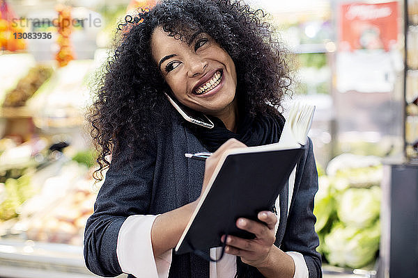 Glückliche Frau telefoniert mit einem Handy  während sie auf dem Markt in ein Buch schreibt
