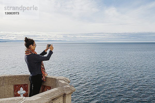 Seitenansicht einer jungen Frau  die das Meer mit einem Smartphone fotografiert  während sie auf einer Gebäudeterrasse gegen den Himmel steht