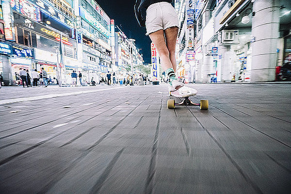 Niedriger Abschnitt einer Frau beim Skateboarden auf einem Fußweg in einer beleuchteten Stadt bei Nacht