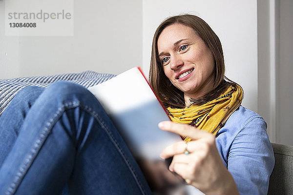 Lächelnde Frau liest Zeitschrift  während sie zu Hause auf dem Sofa sitzt