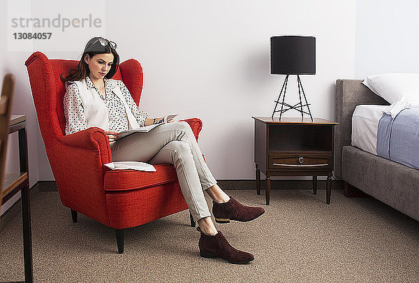 Geschäftsfrau liest Zeitschrift  während sie auf rotem Sessel im Hotelzimmer sitzt
