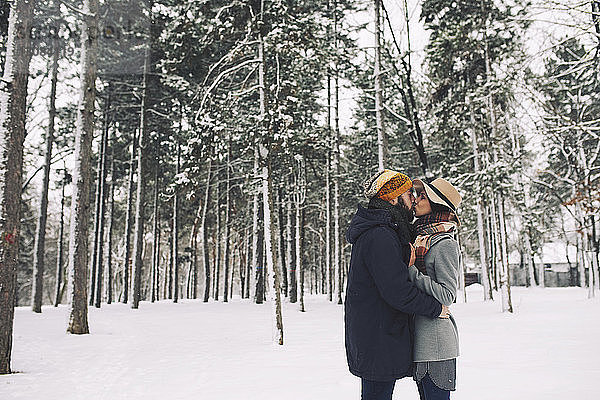 Romantisches Paar küsst sich stehend auf schneebedecktem Feld im Wald