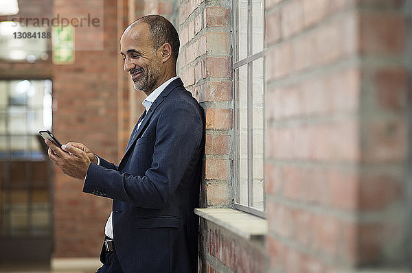 Seitenansicht eines Geschäftsmannes  der ein Smartphone benutzt  während er an einer Ziegelmauer steht