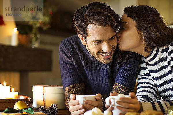 Frau küsst lächelnden Freund  während sie zu Weihnachten Kaffee bei Tisch trinkt