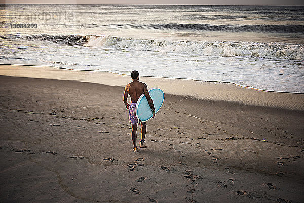 Rückansicht eines Mannes  der ein Surfbrett trägt  während er am Strand auf Sand läuft