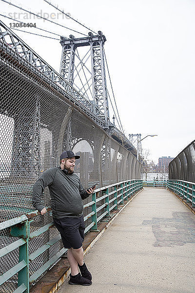Übergewichtiger Mann in voller Länge benutzt ein Smartphone  während er in der Stadt gegen die Williamsburg Bridge steht