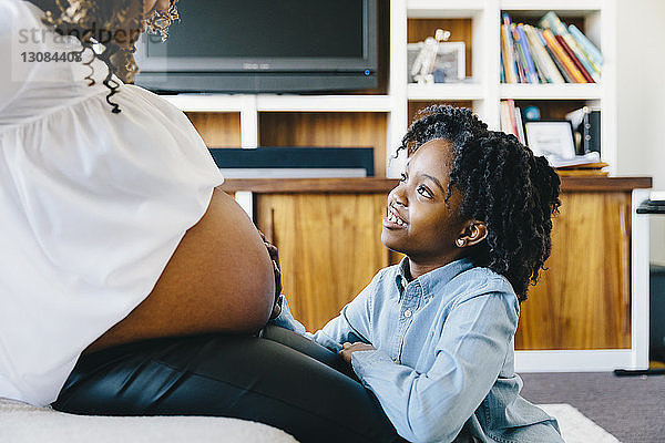 Tochter berührt den Bauch der schwangeren Mutter  die zu Hause auf dem Sofa sitzt