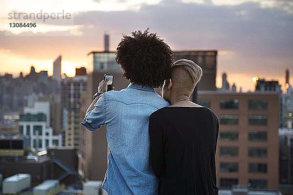 Rückansicht eines Paares  das bei Sonnenuntergang mit einem Mobiltelefon auf dem Dach steht