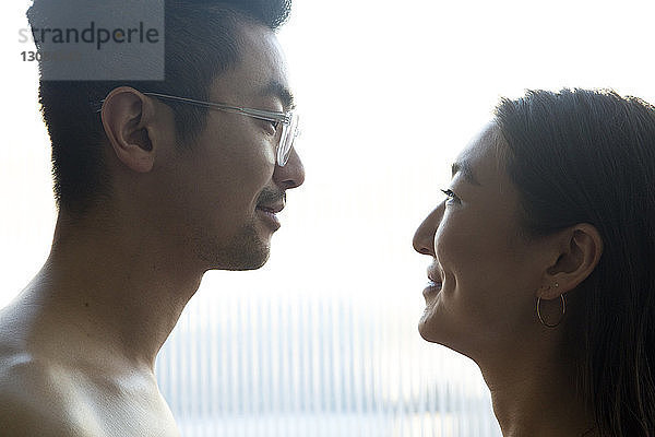 Seitenansicht eines glücklichen Paares  das sich gegenseitig ansieht  während es zu Hause am Fenster steht