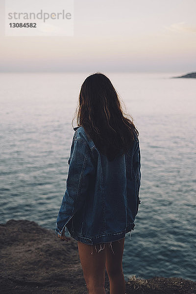 Rückansicht einer Frau  die auf das Meer schaut  während sie bei Sonnenuntergang auf einem Felsen gegen den Himmel steht