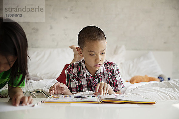 Junge liest Bilderbuch  während er zu Hause neben seiner Schwester im Bett liegt