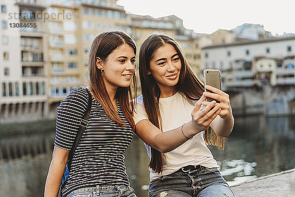 Freunde nehmen Selfie mit dem Handy  während sie auf einer Stützmauer am Fluss Arno in der Stadt sitzen