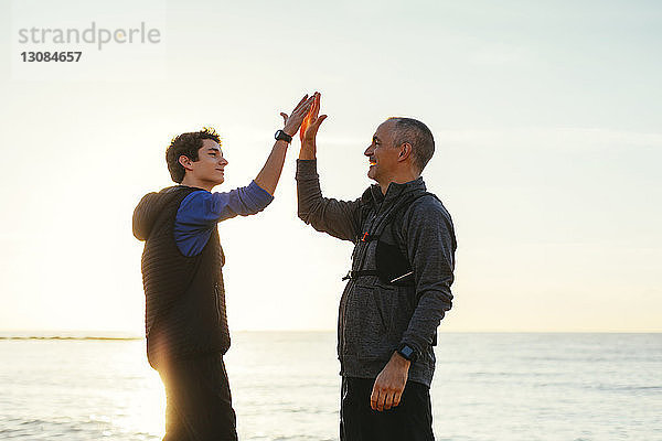 Glückliche Vater und Sohn machen bei Sonnenuntergang ein High-Five gegen Meer und Himmel