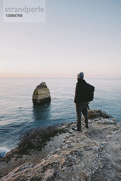 Seitenansicht eines Mannes  der auf das Meer schaut  während er bei Sonnenuntergang auf einer Felsformation gegen den Himmel steht