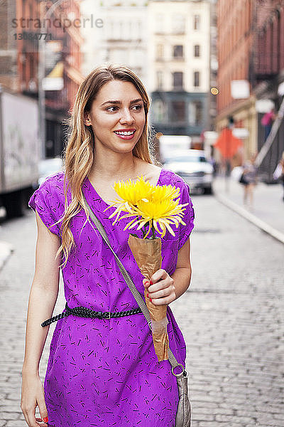 Lächelnde Frau hält Blumenstrauss beim Spaziergang auf gepflasterter Straße in der Stadt