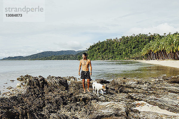 Wanderer ohne Hemd mit Hund auf Felsen stehend gegen den Himmel am Strand