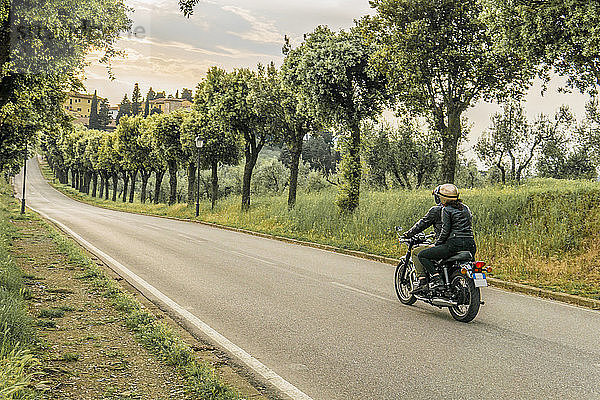 Paar fährt Motorrad auf der Straße zwischen Bäumen bei Sonnenuntergang