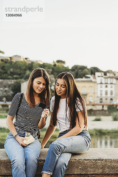 Freunde betrachten ein Sofortbild  während sie auf einer Stützmauer am Arno-Fluss vor klarem Himmel sitzen