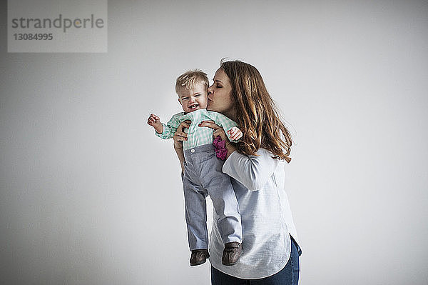 Mutter küsst einen kleinen Jungen  während sie zu Hause an einer weißen Wand steht