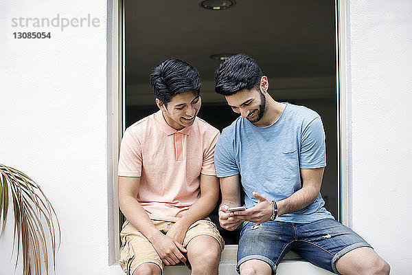 Glückliche männliche Freunde nutzen Smartphone  während sie am Fenster sitzen