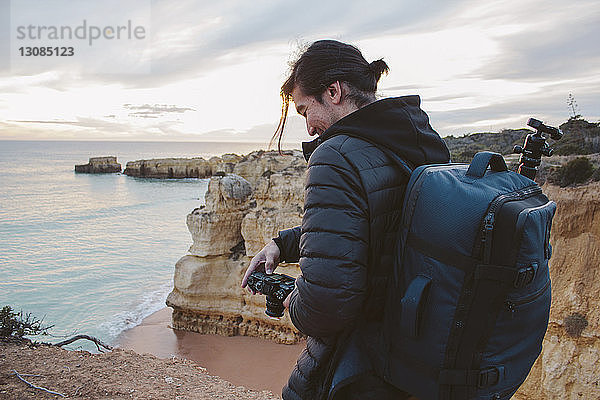 Seitenansicht eines Mannes  der eine Kamera hält  während er auf einer Klippe am Meer vor bewölktem Himmel steht