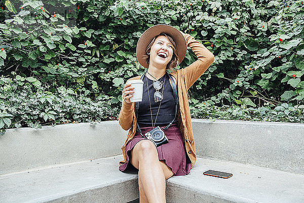 Fröhliche Frau hält Einwegbecher in der Hand  während sie im Park auf einer Bank sitzt