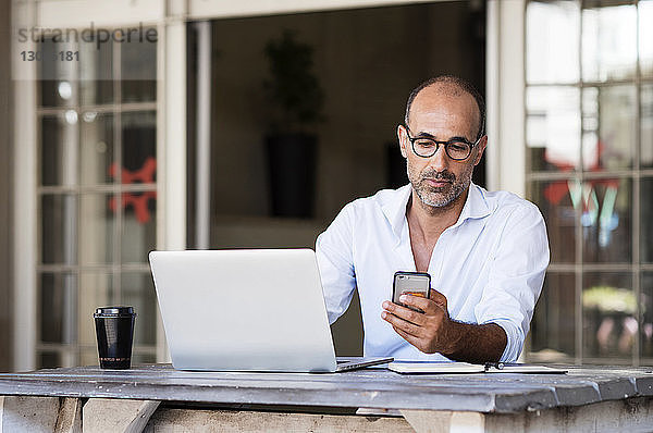 Geschäftsmann benutzt ein Smartphone  während er mit seinem Laptop am Tisch im Büro sitzt