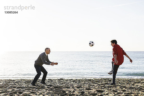 Verspielter Sohn kickt Fussball  während der Vater am Strand gegen den klaren Himmel verteidigt
