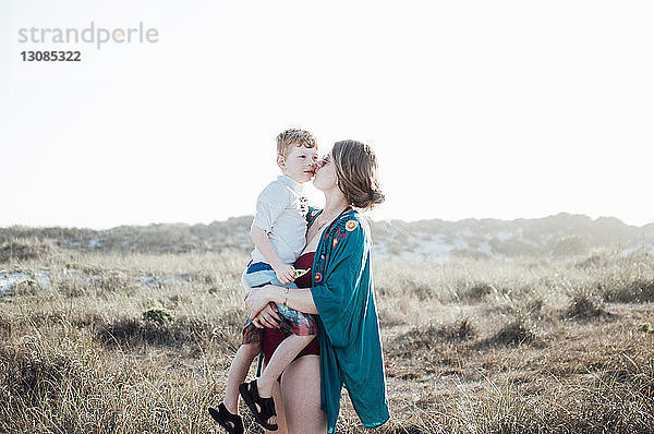 Mutter küsst Sohn  während sie bei strahlendem Sonnenschein auf Grasfeld vor klarem Himmel steht