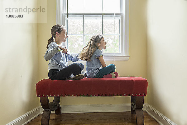 Seitenansicht eines Mädchens  das die Haare seiner Schwester kämmt  während es zu Hause auf einem Sitz am Fenster sitzt