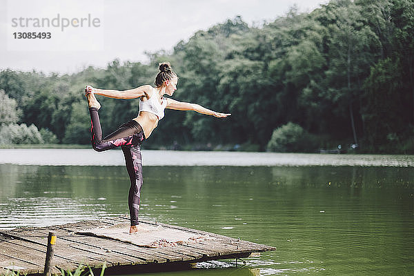 Frau praktiziert Yoga  während sie auf einem Steg über einem See im Wald steht