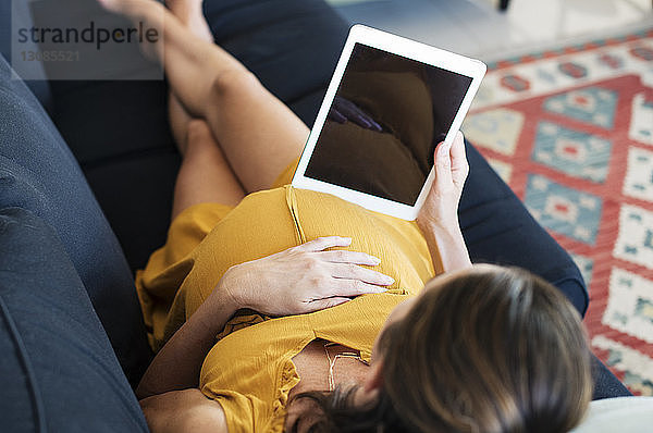 Hochwinkelaufnahme einer schwangeren Frau mit einem Tablet-Computer  während sie zu Hause auf dem Sofa liegt