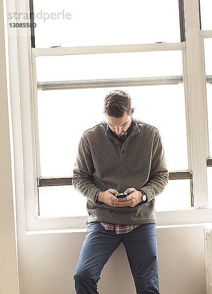 Geschäftsmann benutzt Mobiltelefon  während er sich am Fensterbrett eines hell erleuchteten Büros anlehnt