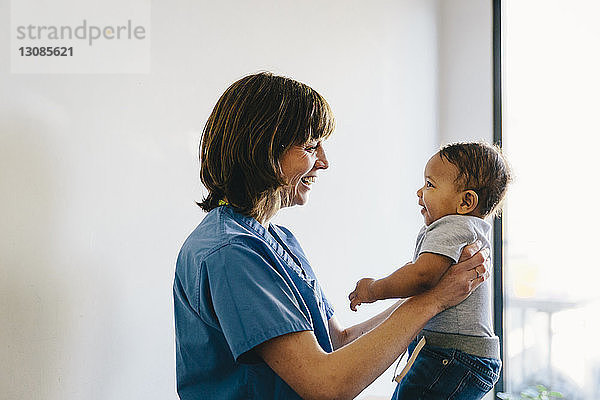 Lächelnde Ärztin  die einen kleinen Jungen trägt  während sie im Krankenhaus am Fenster steht