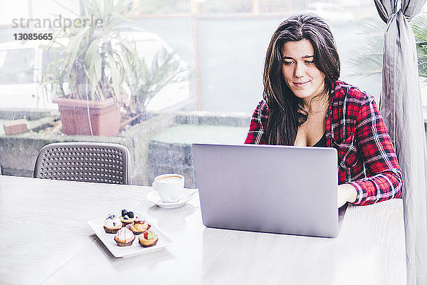 Lächelnde Frau mit Laptop-Computer bei süßem Essen und Kaffee auf dem Tisch im Café