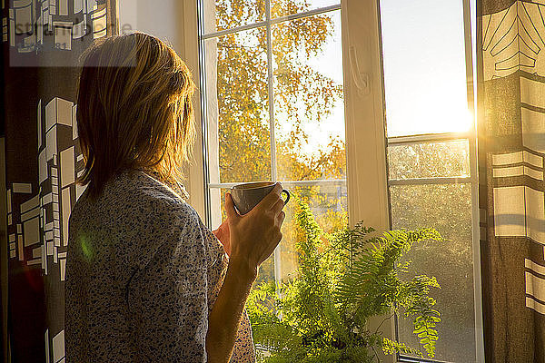 Seitenansicht einer Frau  die eine Kaffeetasse hält  während sie zu Hause am Fenster steht
