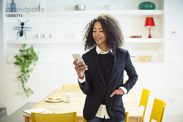 Lächelnde Geschäftsfrau benutzt Mobiltelefon  während sie im Büro am Tisch steht