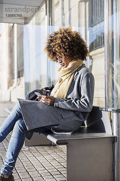 Frau benutzt Mobiltelefon  während sie auf dem Sitz an der Bushaltestelle sitzt